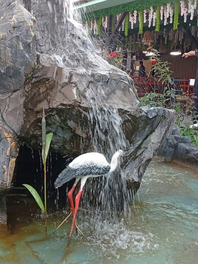 The image depicts a decorative waterfall feature with a statue of a stork in the foreground, standing in shallow water. The stork statue is white and gray with striking red legs. Behind the waterfall, artificial rock formations are visible. The background reveals an outdoor seating area with tables and chairs, decorated with hanging artificial plants and flowers. Several people are present in the background, including one individual pointing towards the scene and others seated at tables. The setting appears to be a cafe or restaurant with an ornamental ambiance, potentially located in Zhibek Zholy, Kazakhstan, given the context provided. The overall scene is brightly lit, suggesting it is daytime.