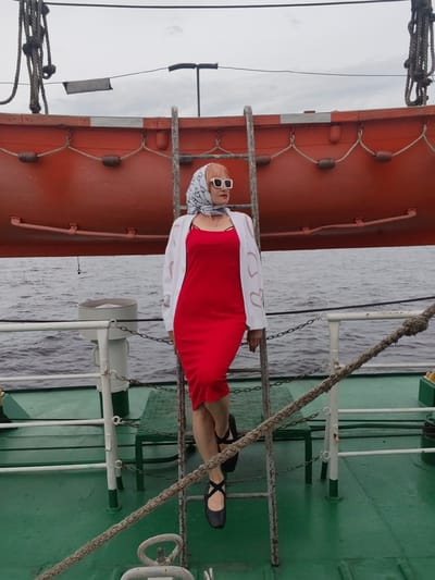 A woman stands on the deck of a ship, leaning against a ladder. She is wearing a red dress, a white cardigan, and a patterned headscarf. Her sunglasses are large and white. The deck is green, and there is a life raft in the background. The sea is visible behind the railing, and the sky is overcast. The scene appears to be a cloudy day, possibly in the morning or afternoon. The woman's pose and attire suggest a casual outing or a posed photograph. There are no visible text elements in the image. The location is Bereg, Russia.