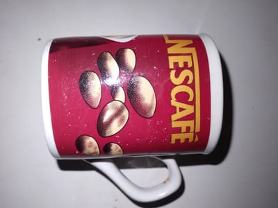 A white ceramic mug with a red wrap-around design featuring coffee beans and the Nescafé logo is lying on its side on a light-colored, textured surface. The mug's handle is visible. The Nescafé logo is in gold lettering. The image is top-down and slightly angled. There are no people or specific geographical markers of Rohati Community of villages, Tajikistan present in this image.