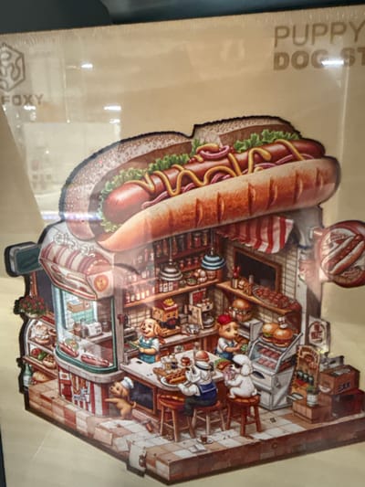 This media file shows a detailed, whimsical illustration of a hot dog stand with anthropomorphic dogs and humans working and interacting. The main subject is a large, illustrated hot dog dominating the left side of the image, complete with bun, sausage, mustard, onions, and lettuce. To the right of the hot dog, a miniature, multi-level hot dog stand is depicted.
The stand features various scenes: a butcher shop area with sausages and meats on display, a serving counter where dog and human characters are interacting, and what appears to be a seating area. Several characters are visible, including dogs dressed in human-like clothing (a chef's hat, a sailor outfit, an apron) and human figures also in attire fitting for a food service establishment. One dog is seated on a stool, appearing to eat. Another dog, wearing a chef's hat, stands near a display case. A human child character is also seen.
Notable details include shelves stocked with food items, a cash register, and signage. The scene is brightly lit, suggesting daytime. The overall atmosphere is cheerful and bustling.
Visible text includes "PUPPY DOG ST" on the left side of the background, partially obscured. On the bottom left, "FOXY" is also visible. A small sign on one of the buildings in the stand reads "BAK". The illustration style is cartoonish and colorful, suggesting a playful and imaginative setting. The media appears to be a printed item, possibly a piece of packaging or a decorative cutout.