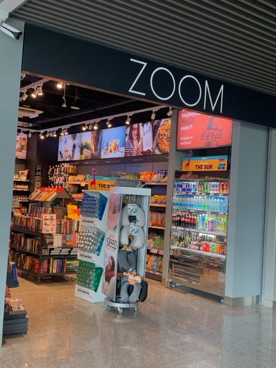 The image shows a retail store named "ZOOM" inside the Pyrzowice Airport, located at 50.471, 19.072. The store is well-lit and features various products on display. There are two large digital signs advertising products. The left sign displays a woman with a smile while the right sign displays a Coca-Cola advertisement. On the left side of the store, there are shelves stacked with books and magazines. In the center, there is a display stand holding various pillows and neck rests. On the right side of the store, there is a refrigerated cooler with rows of beverages including Coca-Cola. The floor is tiled and the walls are painted a dark grey. The store is empty of customers.