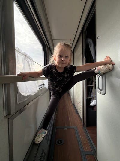This image captures a young girl performing an acrobatic split within a train compartment in Urusovo, Russia. The photo appears to be taken with the camera rotated, giving the impression that the girl is positioned vertically or upside down.
The main subject is a blonde-haired young girl with blue eyes, looking directly at the camera with a playful and confident expression. She is wearing a dark t-shirt with a light floral pattern and dark leggings. On her feet are light-colored Crocs. She is mid-split, with her left leg extended upwards, her left foot braced against an overhead handle or fixture on the train's ceiling/upper bunk structure. Her right leg extends downwards, and her right hand holds onto a metal railing or support bar for balance.
The setting is the interior of a train carriage, likely a sleeping car or couchette, characterized by functional design and compact spaces. Visible features include light-colored ceiling panels, a window to the left showing an overcast sky, and a white sheer curtain hanging near the window. To the right, there are dark wooden-textured wall panels and what appears to be a closed compartment or door. Above the girl's head, an open overhead storage area reveals some folded fabric or luggage. The lighting suggests it is daytime, with soft, diffused light coming through the window. No discernible text is clearly visible. The overall mood is one of lightheartedness and youthful energy during travel.