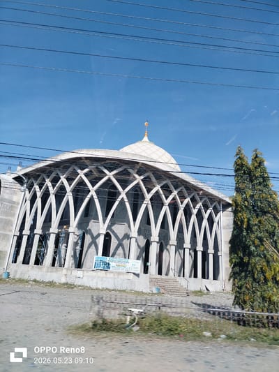 An eye-level shot captures a mosque under construction in Teungoh Baro, Indonesia. The structure features a white dome with a spire, supported by a series of arched white concrete frames. A tree stands to the right of the mosque. The building is surrounded by what appears to be a construction zone. Overhead power lines cross the sky. The sky is blue with some light clouds. A sign is posted at the base of the building. The image was taken on May 23, 2026, at 09:10 AM.