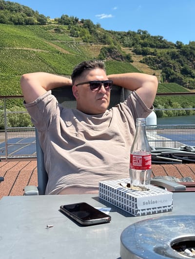 A man sits in a chair on a deck with his hands behind his head, wearing sunglasses and a tan t-shirt. In front of him on a table is a smartphone, a box of tissues labeled "satino HiLine", an empty glass bottle with a red label, and an ashtray containing cigarette butts. The background shows a steep hillside covered in vineyards and a body of water. The scene is set in Piesport, Germany, a region known for its vineyards and Moselle River wine production.