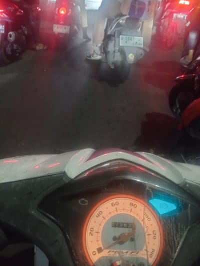 The image shows a close-up view of a motorcycle's dashboard, specifically the speedometer and some indicator lights. The speedometer dial is orange and white, with markings up to 140. A digital display above the speedometer reads "37363". To the right of the speedometer, a blue indicator light, possibly for headlights, is illuminated. The dashboard itself is primarily black plastic, with some white and pink accents on the surrounding fairing. The background of the image is dark and appears to be an outdoor setting, possibly at night, with hints of other motorcycles and faint red lights, suggesting a street or parking area. The overall scene is dimly lit. No people are clearly visible in the foreground, but the presence of other vehicles and the context of a dashboard suggest a transportation-related environment. The specific location, Calukan RT 1 RW 5 Keboansikep, Indonesia, is noted but not visually depicted in the provided image.