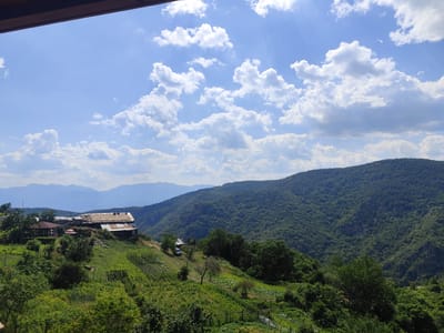 A view from a wooden balcony looks out over a lush, green mountainous landscape. In the foreground, terraced fields are visible with various crops and vegetation. Several rustic structures, appearing to be houses or farm buildings, are situated on a hillside. These structures have weathered roofs and some appear to be in disrepair. A dark-colored vehicle is parked near one of the buildings.
The middle ground is dominated by rolling green hills covered in dense forestation. The background consists of several layers of hazy blue mountains extending to the horizon. The sky is bright blue with scattered fluffy white clouds, and sunbeams are visible filtering through them. The overall scene depicts a rural, mountainous area, likely in the village of Leshten, Bulgaria, characterized by its natural beauty and rustic dwellings.