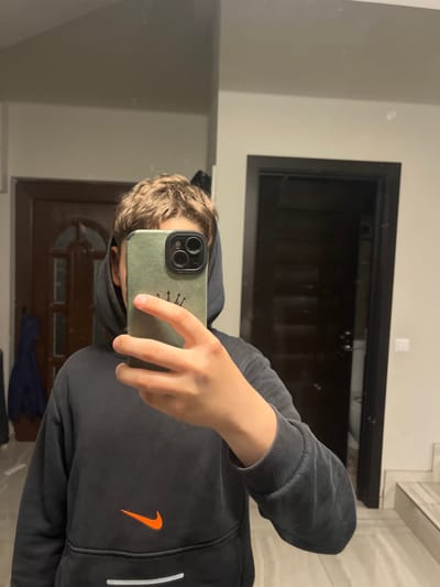 A mirror selfie captures a young individual, likely male, with short, wavy light brown hair, holding a smartphone. The phone is in their right hand and encased in a sage green cover featuring a black triple-lens camera module and a small gold crown logo. The person is wearing a black hoodie, prominent with an orange Nike swoosh logo on the front and a white stripe visible along the side. The ribbed cuff of the hoodie's sleeve is also discernible.
The reflection in the mirror reveals an indoor setting with light-colored, possibly off-white or light gray, walls. Two dark-framed doorways are visible in the background; one is dark and angular, appearing directly behind the person's head, while another, seemingly a wooden door with panelling, is reflected lower down. The lighting suggests an indoor environment, likely artificial. The scene is located in Cherni Vrah, Bulgaria.