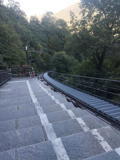 This media file depicts a steep, outdoor staircase made of granite, with white stripes painted on some of the steps. To the right of the staircase, a funicular track runs parallel to it, constructed with metal grating for the steps. The track curves to the right and disappears into the background. A metal railing lines the right side of the funicular track and also borders the top of the staircase on the left.
In the background, a lush, green forested mountainside rises steeply. The trees are dense and varied, with some taller trees visible amongst the undergrowth. The sky above is hazy and bright, suggesting either early morning or late afternoon light, possibly with some mist or fog.
At the top of the staircase, near the railing, there are two red and white striped stanchions connected by a red and white striped tape, indicating a temporary closure or barrier. Behind these, some pink flowers are visible on a bush. A dark metal pole with a white device at the top, possibly a light or sensor, stands near the railing further up the staircase.
The scene appears serene and tranquil, with no people visible. The overall impression is one of a scenic and slightly rugged outdoor environment, likely a tourist location or a path leading to a vantage point. The location context of Orselina, Switzerland, suggests a mountainous or hillside setting, which aligns with the visual cues in the image.