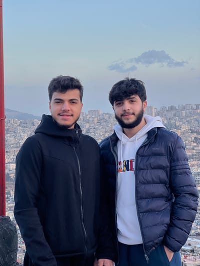 The image displays two young adult males standing outdoors from an elevated perspective overlooking the city of Qudsaiya, Syria. The individual on the left has short, dark hair and a light beard, wearing a black zip-up hooded jacket. He is looking forward with a slight smile. The individual on the right has dark, curly hair and a darker beard, wearing a white hooded sweatshirt with a colorful "AND" graphic under a dark blue puffer jacket. He is also looking forward with a slight smile.

Behind the subjects, the urban landscape of Qudsaiya stretches across the frame, characterized by numerous densely packed, light-colored residential and commercial buildings. A distant mountainous terrain is visible on the far left horizon. The sky is a clear light blue with a single wispy cloud present in the upper right quadrant. A red, vertical pole or structure is partially visible on the extreme left edge of the image. The lighting suggests late afternoon or early morning conditions.