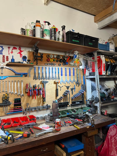 A cluttered workbench holds an array of tools: wrenches, pliers, hammers, screwdrivers, and specialized equipment, neatly organized on a wooden panel. Cans, crates, and boxes overflow on shelves above, while miscellaneous items occupy the right side of the workbench, including a Coca-Cola can. A dark cabinet sits beside the workbench.