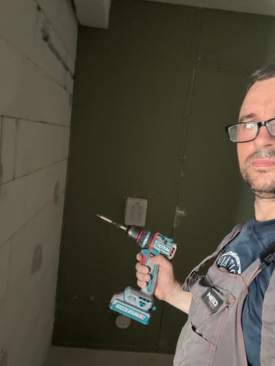 In this image, a person is seen holding a cordless drill. The person is wearing a dark blue shirt and grey overalls with orange stitching, featuring a "NEO TOOLS" logo patch. The drill itself is teal and white, with "TOTAL" branding clearly visible on its body, along with "20V" and other model identifiers. The person is wearing glasses and has facial hair, and their gaze appears to be directed downwards or at the drill. The background consists of a dark olive green wall with faint lines suggesting paneling or joints, and possibly some small indentations or marks. Below the wall is a light-colored floor, likely made of wood or a similar material, with visible seams between planks. The lighting is dim, suggesting an indoor environment, possibly during the evening or in a poorly lit area. There are no clear indications of weather or time of day beyond the ambient light. The overall scene suggests a DIY or construction/renovation context, with the person engaged in an activity that requires the use of the drill. The location is identified as Sušćepan, Montenegro, though this context is not visually evident within the frame itself.