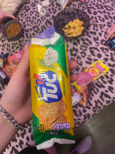 A hand, adorned with a chunky silver bracelet, holds a yellow and green package of TUC crackers. The crackers have a visible flavor of "Smetana and Onion," indicated by an image of sour cream and green onions on the packaging. The background is a table covered with a leopard-print tablecloth. Various items are scattered on the table, including bowls of snacks and a pink and yellow lollipop wrapper. A glimpse of a phone with a picture on the screen can be seen on the right. The scene appears to be a casual gathering, possibly a party or a get-together. The specific location of Zmeinyi, Russia is not discernable from the image.