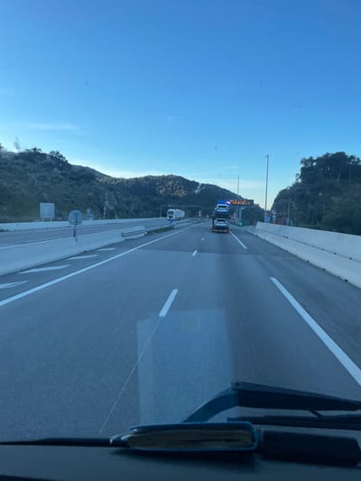 The image depicts a multi-lane highway environment near Le Perthus, France, viewed from within a vehicle. The sky is clear blue. Hilly terrain, densely covered with green vegetation and some rocky outcrops, flanks both sides of the highway.
In the immediate foreground, a vehicle's dashboard and two windshield wipers are visible. The asphalt road features white dashed and solid lane markings. White chevron markings are present on a merge lane to the left. Concrete barriers separate the main carriageway from the median and shoulders.
Vehicular traffic includes a white car carrier truck occupying the central lane, transporting four vehicles: two white cars are stacked on its upper deck, and two darker-colored vehicles are positioned on its lower deck. Further ahead in the left lane, a white semi-trailer truck is also in motion.
Overhead gantries with visible fixtures span the highway. On the right side, an electronic variable message sign displays illuminated yellow text, with the word "ALERT" clearly discernible. A small structure is situated roadside to the right. Distant mountainous terrain forms the horizon.