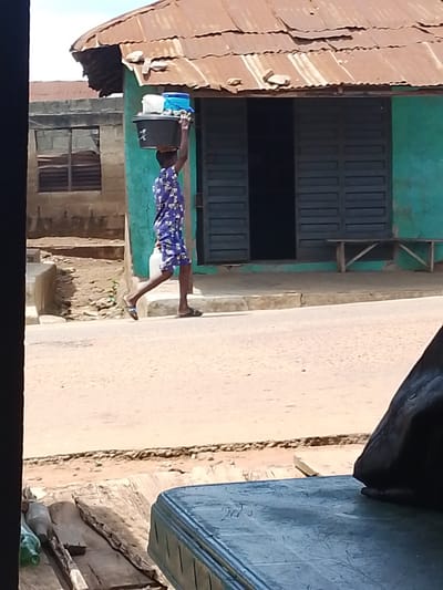 A person is walking on a dirt road in front of a building with a green painted facade and a corrugated iron roof. The person is carrying a large black basin filled with items on their head. The basin contains several blue and white containers. The person is wearing a blue and white patterned dress and flip-flops. The building has a dark doorway and a wooden bench outside. A concrete structure and a building with a barred window are visible to the left of the main building. The scene appears to be set in a sunny, outdoor environment, likely during the daytime. The overall setting suggests a village or town in Nigeria, specifically Iseyin, based on the provided context. The image conveys a sense of everyday life and labor.