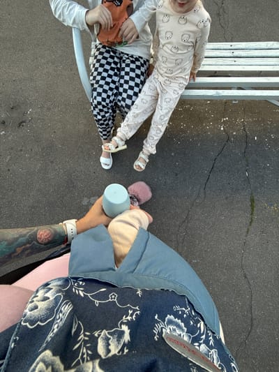 Two children are sitting on a white bench outdoors. The child on the left is wearing a white long-sleeved shirt with an orange graphic on the front and black and white checkered pants. They are wearing white sandals. The child on the right is wearing a light pink long-sleeved pajama set with a pattern of cartoon bears. They are also wearing white sandals. In the foreground, a person's arm with a tattoo is holding a light blue cup and a pink fluffy object. This person is wearing a blue garment with a floral pattern. The ground appears to be asphalt with some cracks. This scene is located in the city of Uzunovo, Russia.