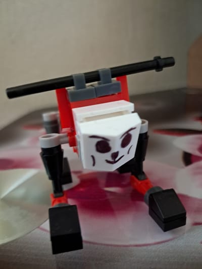 The image presents a custom-built figure, resembling a character constructed from building blocks, likely LEGO or a similar brand. The figure is oriented horizontally, against a softly blurred background.
The "head" of the figure is a white rectangular block, on which a face has been drawn with dark brown or black marker. This face features circular eyes, a small nose, and a downturned mouth, giving the character a somewhat melancholic or "oof" expression.
Below the head, the "body" is primarily composed of red blocks. Extending from the body are four limbs, made from black rod pieces, which terminate in flat, black rectangular blocks, acting as hands and feet. These limbs are connected to the main body via small red and grey joint elements. On the left side of the figure's body, a long black rod is attached vertically using two grey clip pieces, possibly representing a staff, weapon, or handle.
The background is indistinct, displaying soft blurs of white, pink, and reddish tones, suggesting an indoor setting with patterned elements behind the figure. No activity is depicted; the figure is static, observed as a photographic subject. There is no visible text in the image.