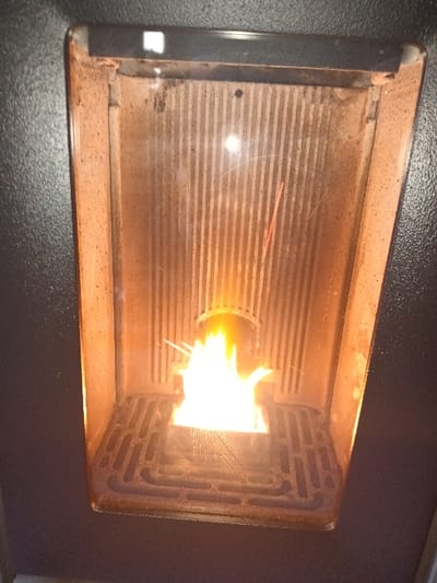 The image displays a close-up view of the interior of an active pellet stove's combustion chamber, situated in Libramont, Belgium. A vibrant yellow and orange flame emanates from a square metal burn pot positioned on a dark, patterned cast-iron grate at the bottom of the firebox. The vertical interior walls are corrugated and display discoloration and soot consistent with heat exposure. A dark horizontal panel across the top interior features a small white rectangular indicator light on its left side. The stove's visible exterior frame is dark-colored. No individuals or other objects are visible.