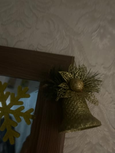The scene depicts a close-up, upward-angled view of a golden Christmas bell ornament attached to the upper right corner of a dark brown wooden frame, possibly a mirror or picture frame, located in Dmitriyevskaya, Russia. The bell is covered in glitter and features a decorative top comprising intricate gold mesh leaves or petals, a central golden glittered sphere, and dark green faux pine needles. To the left of the bell, a segment of a reflective surface, likely a mirror, shows a blurred yellow snowflake decoration and indistinct light reflections. The background consists of a light-colored wall with an embossed, swirling pattern, indicating textured wallpaper or plaster. The overall lighting is dim and ambient, illuminating the decorative elements and wall texture. No people or active interactions are present in the frame.