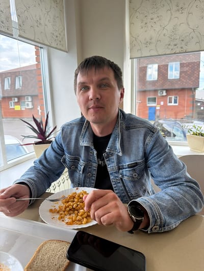 A man in a blue denim jacket is at a table. He is holding a fork and a plate of macaroni with meat. Beside the plate is a smartphone and a slice of bread. The man is wearing a watch on his left wrist. The scene is likely inside a building as there is a window behind the man. Through the window, there is a view of other buildings, suggesting an urban area. There is no available data to verify the location of Kulaevo, Russia from this image. The picture does not provide any information on this location.