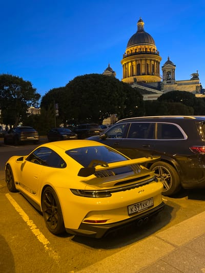 A yellow Porsche GT3 is parked in a lot in front of a large domed building, possibly St. Isaac's Cathedral in Saint Petersburg, Russia, as indicated by the license plate reading "K391KK 77 RUS". The car is positioned in the foreground, its rear end facing the viewer. Behind it, a dark-colored SUV is parked. The sky is a deep blue, suggesting it is either early morning or late evening. Several trees are visible around the parking area. The lighting is low, with the dome and some architectural elements of the building illuminated.