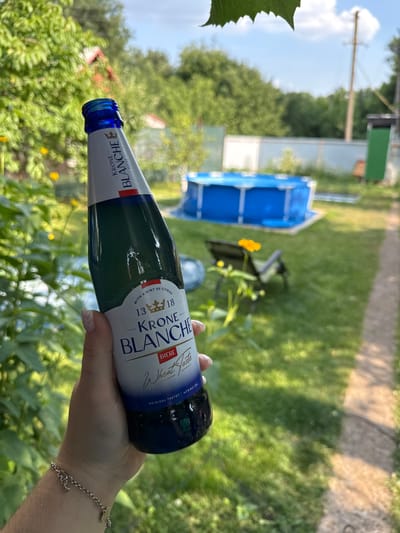 A person's hand, adorned with a delicate bracelet, holds a glass bottle of Krone Blanche Biere. The bottle, which appears to contain a pale liquid, has a label with text including "KRONE BLANCHE BIERE," "Wheat Fasts," and "4.5% ALC/VOL." The label also mentions "WITH A HINT OF CITRUS." The background suggests an outdoor setting, likely a backyard, with lush green grass and trees. A blue inflatable pool is visible in the distance, along with a path or walkway. The lighting suggests it is daytime, possibly a sunny day given the bright green foliage. The image is oriented sideways, with the bottle held horizontally.