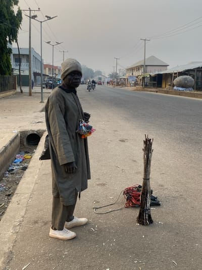 A man wearing a suit and a cap stands beside a street drain. He holds colorful items in his hand. Next to him is a bundle of wooden sticks tied together, with a set of red cords attached. The scene is set in a street in daylight. Buildings and utility poles are visible in the background. There are motorcycles driving in the distance on the street.