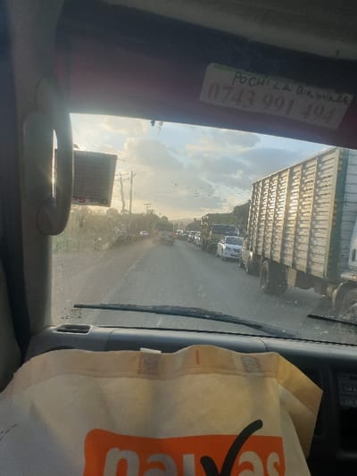 The image displays an exterior view from the interior of a vehicle, positioned on a dusty road in Mumbuni North ward, Kenya. The perspective is through the windshield, which exhibits some dust or smudges. The scene is lit by a low-angle sun, indicating either early morning or late afternoon, casting a golden light and creating a slightly hazy atmosphere with scattered clouds in the sky.
Inside the vehicle, a portion of the dark-colored dashboard is visible, along with a single windshield wiper blade resting at the base of the glass. A light-colored fabric bag, marked with orange text partially discernible as "naivas" and a black checkmark, rests on the dashboard. The red interior roof lining is visible at the top, bearing a white rectangular sticker with handwritten text "POCHILA BIRSHAA" and a phone number "0743 991 494". To the left, a white tag, partially reading "ONE SE", hangs from an interior grab handle.
Outside, a substantial line of vehicles, including cars and trucks, extends into the distance, suggesting a traffic jam or slow movement. Immediately adjacent to the right side of the viewer's vehicle is a large, dark-colored cargo truck with a ribbed side, reflecting the direct sunlight. Further down the road, a white sedan and additional trucks are visible in the queue. On the left side of the road, a utility pole with multiple cross-arms and power lines is present. The road surface appears to be unpaved or gravel, contributing to the dusty environment. Distant vegetation is visible along the horizon. No individuals are discernible within the frame.