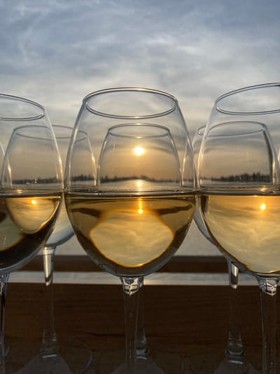 The image depicts a row of wine glasses, some filled with a yellow liquid, likely wine. The glasses are arranged on a surface, possibly a table or ledge, with an outdoor background visible through the clear sections of the glasses. The background appears to be a sky with clouds. The light is bright, suggesting daylight. The reflections in the liquid-filled portions of the glasses suggest a sunset or sunrise scene. The focus is on the glasses, with the background somewhat blurred. There is no indication of this image being located in the city of Aramil, Russia.