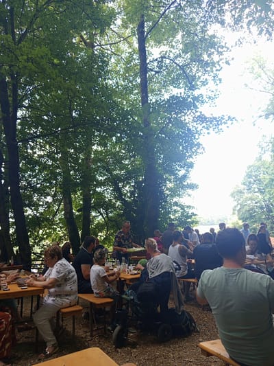This image depicts an outdoor gathering of people, possibly a picnic or a community event, set in a wooded area with dense foliage overhead. The scene appears to be informal, with individuals seated at wooden tables and benches.
In the foreground, a woman with glasses and a white floral patterned shirt sits at a table, appearing to be engaged in an activity. To her right, a young boy in a camouflage-patterned hat and shorts sits on a bench. In the midground, several people are seen interacting at tables, some are eating or drinking. A man in a floral shirt is standing and appears to be serving or attending to a table. The background shows more trees and a bright sky, suggesting it's daytime. The ground is covered with gravel and fallen leaves.
Given the context of Azannes-et-Soumazannes, France, this could be a local festivity, a family reunion, or a casual social gathering in a park or forested area common in the region. The presence of multiple tables and groups of people suggests a communal event. The overall atmosphere is relaxed and social.