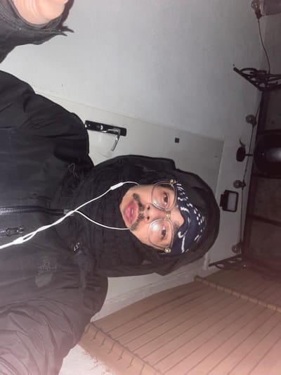 A person wearing glasses, a navy bandana, and a black jacket with a scarf pulled up around their neck takes a selfie. They also have white wired earphones in. The background includes a white wall and door with a silver handle. Overhead are various lighting fixtures and decorative wrought iron. The photo appears to be taken at night or in a dimly lit area.