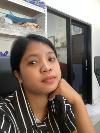 A person with dark hair and brown skin is visible in this indoor selfie. They are wearing a black and white striped shirt. The background features a dark-framed window and a shelf with items wrapped in plastic. Based on the location provided, the photo was likely taken in Negara, Indonesia.