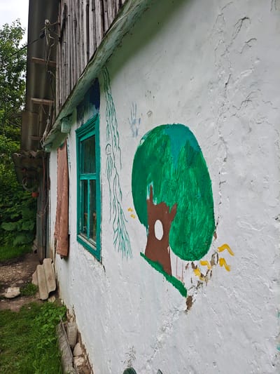 This image captures a section of a weathered, white-painted building, likely a rural house, in Akhmetovskaya, Russia. The wall is adorned with naive, colorful paintings. A prominent feature is a large, stylized green tree with a brown trunk and a white circular opening. To the left of the tree, a drawing of a handprint is visible in blue, and a green vine-like motif with thin tendrils extends downwards. To the right of the tree, there are a few yellow brushstrokes that could represent sunlight or stylized flowers. The building itself shows signs of age, with peeling paint and cracks in the plaster, particularly at the base where some debris and greenery are visible.
A bright turquoise window with multiple panes is set into the wall. To the left of the window, a piece of rough, brown fabric is draped, possibly a curtain or a tarp. In the background, to the left of the building, a dense green foliage of trees and bushes can be seen. The upper part of the building has a wooden, vertical siding and a slightly slanted roof with a visible gutter. Electrical wires and insulators are attached to the wooden siding, suggesting functional utilities. The overall impression is one of a simple, perhaps rustic, dwelling decorated with folk art. The lighting suggests it is daytime, with overcast or diffused light. There are no people visible in the image. No discernible text is present other than what might be part of the painting, which is too stylized to read.