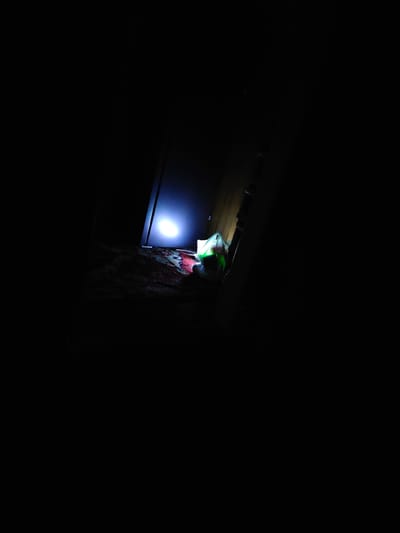 An illuminated object, possibly a bag with a green light emanating from it, rests on a patterned carpet on the floor. Behind the object, a bright white light emanates from an unseen source, casting a glow on a dark vertical surface, possibly a door or wall. The environment is predominantly dark, with only the illuminated areas being clearly visible. The scene appears to be indoors.