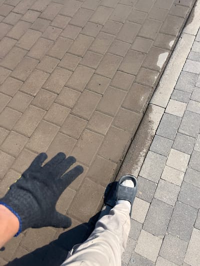 The image shows a close-up of a person's lower legs and feet, as well as a gloved hand, resting on a paved surface. The surface is divided into two sections by a curb, with lighter colored pavers on the upper right and darker, rectangular pavers on the lower left. There are wet patches on the pavers and curb, suggesting recent rain or cleaning. The person is wearing a black sandal on their right foot, a white sock, and light-colored trousers. A black glove with blue trim is visible on the left hand, casting a shadow on the pavers. The scene appears to be outdoors, likely in a park or a residential area given the paving stones. The lighting suggests it is daytime, with the sun casting shadows. The overall impression is casual and practical, possibly indicating yard work or simply a moment of rest.
