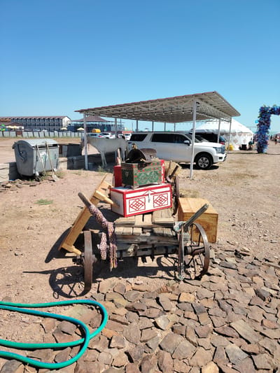 In the city of Torangylyk, Kazakhstan, an outdoor scene unfolds under a clear blue sky. A weathered wooden cart, adorned with patterned fabric and metal accents, sits prominently in the foreground. On the cart are two decorative chests, one red and white with intricate geometric patterns, and another smaller green chest with ornate detailing. To the left of the cart, a green garden hose lies coiled on a bed of flat, reddish-brown stones. In the background, a white horse stands near a modern white SUV parked under a white-canopied structure. Further back, a long, multi-story building with numerous windows and balconies is visible, alongside other structures and a large white tent-like building. To the far right, a decorative archway draped in blue flowers adds a touch of festivity to the scene. The ground is a mix of gravel and dirt, suggesting a dry, possibly arid environment.
