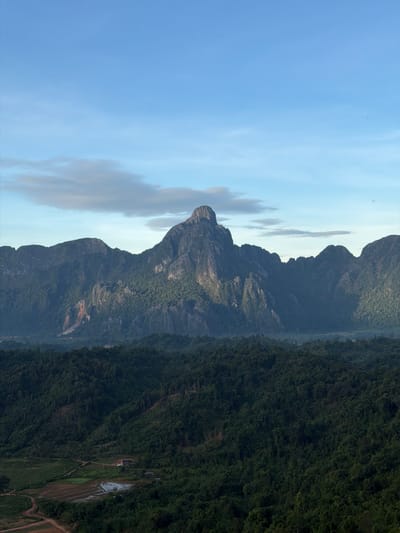 This is a vertical, wide-angle photograph capturing a mountain range and a forest. The sky is clear blue with a few wispy clouds near the horizon. The mountains are jagged and rocky, covered in lush green vegetation. In the foreground, a dense forest stretches across rolling hills. There is a small clearing in the forest with a few buildings, including what appears to be a house with a red roof, and some agricultural fields. The overall atmosphere is serene and natural. The location is identified as Naka, Laos.