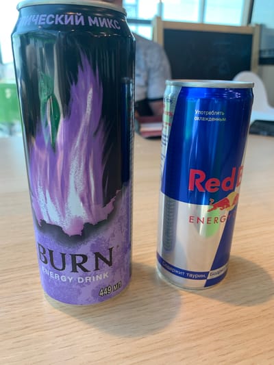 Two energy drink cans are on a light-colored wooden surface. The can on the left is predominantly black with purple and green flame-like graphics and the text "BURN ENERGY DRINK" in purple and white, along with "449 мл" in white. The can on the right is blue with a silver bottom portion, featuring the Red Bull logo and the text "Red Bull ENERGY" in red. Russian text is visible on both cans. In the background, a person wearing a grey shirt is partially visible, and a dark surface with white framing is behind them, possibly a blackboard. The environment appears to be indoors, with bright lighting from windows in the upper background. There is no indication of the specific location in Bol'shie Kaibitsy, Russia.