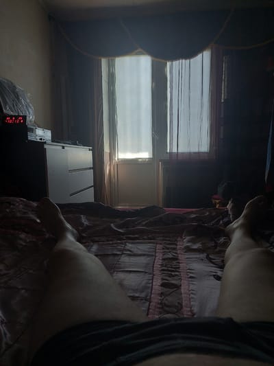 The image shows a dimly lit bedroom with a person lying on a bed, facing away from the camera. The view is from the perspective of the person lying down, with their legs and feet visible in the foreground. The bed is covered with a patterned comforter. In the background, there is a dresser with a digital clock displaying "10:13". A window with sheer curtains is visible, letting in some natural light. The overall atmosphere is one of domestic intimacy and quiet. The location is specified as Cimișlia, Moldova, though no specific landmarks confirm this.