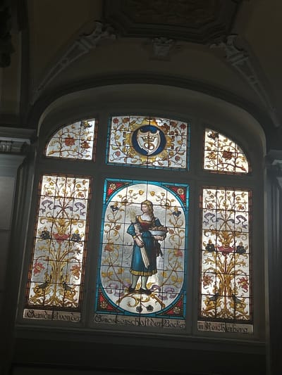 This image showcases a large, elaborate stained glass window within a grand interior setting in Liberec, Czechia. Bright light streams through the window, illuminating its intricate details and vibrant colors.
The window is divided into several sections. The prominent central panel features a female figure, possibly a saint or an allegorical representation, dressed in a blue gown and holding a book or scroll. She is surrounded by delicate floral and foliate patterns in shades of gold, green, and red, bordered by blue and red glass. To her left, another panel contains a circular motif with what appears to be an intricate monogram or symbol, also encircled by ornate floral designs.
Smaller panels above, below, and to the top left of the main sections are filled with decorative floral and organic patterns, primarily in gold, red, and purple against light backgrounds.
The dark frame of the window contrasts with the glowing glass. Text is visible vertically inscribed on the right side of the window frame. The upper inscription reads "in Reichenberg.", identifying the location by its historical German name. Below it, the text reads "Genossenschaft der Bäcker" (Bakers' Cooperative) and "Gewidmet von der" (Dedicated by the), suggesting the window was a gift or commission from a bakers' guild.
The surrounding architecture includes a gracefully curved archway and decorative corbels on the wall, contributing to the historical and ornate ambiance of the space. The scene depicts a richly decorated interior, likely part of a significant building in Liberec. The time of day appears to be daytime, as indicated by the light through the stained glass.