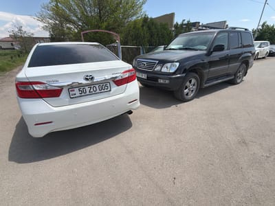 The image shows two parked cars on an asphalt surface in Dimitrov, Armenia. A white Toyota Camry with Armenian license plate "50 ZO 005" is parked to the left, with its rear facing the viewer. Beside it is a black Lexus LX470 SUV with a Russian license plate "777 PP 70" oriented with its right front at the viewer. In the background are trees, a building, and a chain-link fence. Additional vehicles are partially visible further behind the SUV. The weather appears sunny and clear.