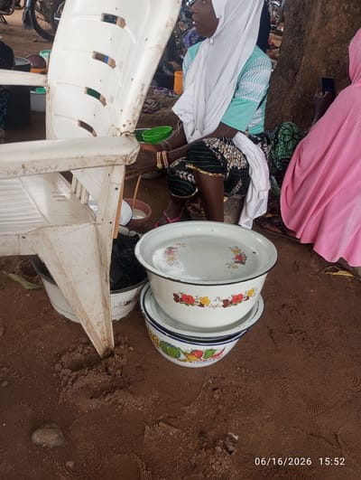 A medium shot shows a group of people sitting on the ground outdoors in Dokan Tofa, Nigeria. A white plastic chair is in the foreground, tilted to the left. Underneath the chair, a large white enameled basin with a floral pattern is visible, containing a black garbage bag. Stacked on top of this basin are two more enameled basins, one with a floral pattern and the other with a fruit and vegetable pattern. To the right of the basins, a woman wearing a pink hijab and a pink garment is sitting and looking at her phone. To the left of her, a woman wearing a white hijab and a turquoise top with white stripes is sitting, with her hands clasped in her lap. The ground is covered in reddish-brown soil. A tree trunk is visible behind the people. The date and time stamp "06/16/2026 15:52" is in the bottom right corner.