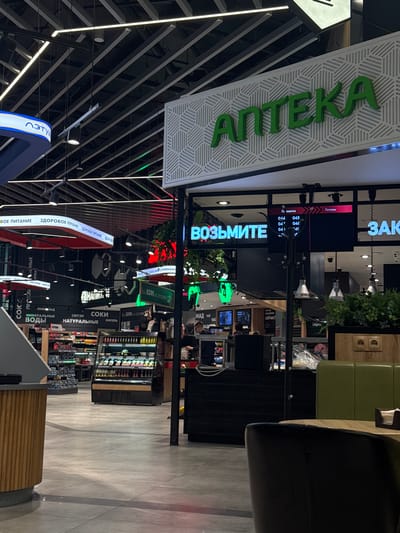 The image shows the interior of a retail establishment, possibly a supermarket or shopping center, in what is likely the city of Vorsha, Russia, given the language on the signs.
**Subjects/Objects:**
* Signage in Russian: includes "АПТЕКА" (pharmacy), "ВОЗЬМИТЕ" (take), "ЗДОРОВОЕ ПИТАНИЕ" (healthy food), "СОКИ" (juices), "НАТУРАЛЬНЫЕ" (natural), "ВОДЫ" (waters), "ЛИМОНАД" (lemonade), etc., suggesting various departments or product categories.
* Refrigerated shelves with various beverages and food items.
* Ceiling with intricate patterns and lighting fixtures.
* A digital screen displaying order numbers or cooking times.
* A counter or service area.
* Partial view of seating (chairs/sofa).
* Other customers and staff members.
**Actions/Interactions:**
* People shopping or browsing.
* Possible interaction between customers and staff.
**Environment:**
* Modern interior design with a clean and well-lit environment.
* Flooring appears to be tile or polished concrete.
* Commercial space with signs directing customers.
**Additional Notes:**
* The presence of both a "pharmacy" sign and food items suggests a combined store or a supermarket with a pharmacy section.
* The signs point toward a commercial environment.