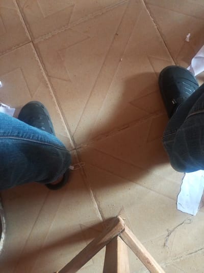 The image shows a close-up view of two people's legs wearing blue jeans and black slip-on shoes, standing on a tiled floor. The tiles are large and appear to be a light brown or beige color, with a pattern of raised geometric shapes. There are pieces of white paper scattered on the floor, and some loose threads. In the lower right of the image, a wooden frame or structure is visible. The scene appears to be indoors, possibly in a home or workshop. The image's composition focuses on the lower bodies of the individuals and the immediate surroundings.