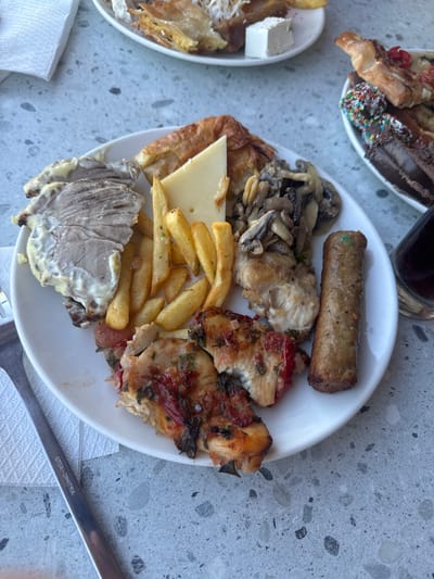 A plate of food sits on a gray, speckled surface, likely a table. The plate is full of various dishes, suggesting a meal, possibly a buffet or a large shared plate.

The main subject is the food itself, which includes a long, grilled sausage with green flecks, a pile of sautéed mushrooms, pieces of baked chicken or poultry topped with red sauce and herbs, a slice of cheese, and a serving of french fries. There are also some pastry-like items and a slice of roasted meat covered in a creamy sauce. In the background, another plate holds a selection of sweet pastries, some topped with colorful sprinkles.

The scene is a close-up, top-down view of the food, with a hint of a glass of dark liquid (possibly soda or wine) and a fork to the right. The lighting suggests it could be daytime, though the angle and overhead view don't provide clear cues about time of day or weather. The presence of multiple dishes and the casual arrangement imply a relaxed dining setting.

Visible text on the fork reads "GTSA-DIMITRA". The location context suggests this meal is in Agios Nikolaos Municipal Unit, Greece. No people are visible in the frame. The overall impression is of a hearty and varied meal, presented in a casual, possibly outdoor or cafeteria-style setting.