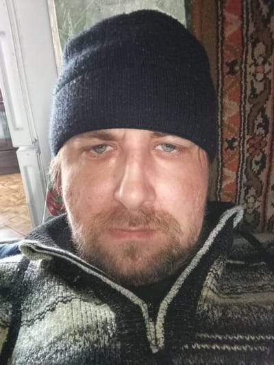 A Caucasian male with light-blue eyes, a short, light-brown beard, and wearing a dark knit cap and a black-and-white patterned zip-up jacket is facing the camera. The background includes a white door frame and a patterned tapestry. The image location is Parabel, Russia.