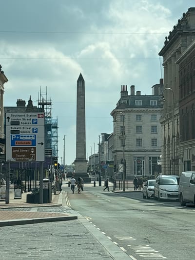 The image depicts a street scene in Southport, United Kingdom. In the foreground, a sign indicates directions to Southport Station, London Street parking spaces, Tulketh Street parking spaces, Esplanade, and Lord Street Tourist Information. Scaffolding is visible behind the sign. Further down the street is a tall, gray obelisk. People are walking on the street, and cars are parked on the side. The sky is overcast.