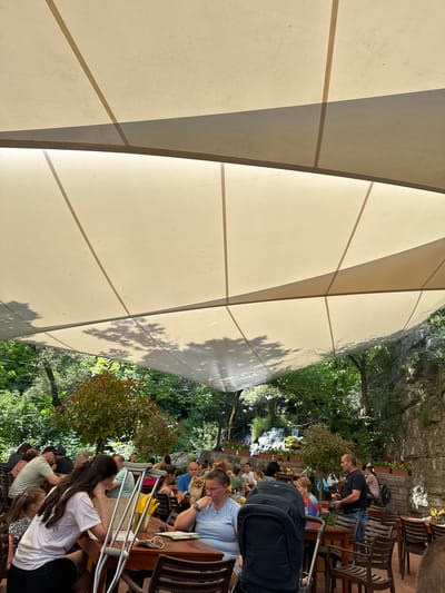 The image depicts an outdoor dining area in Milanovo, Bulgaria. Several individuals are seated at tables, some eating and conversing, positioned under a large, angular, canvas awning. Vegetation, including trees, is visible in the background, along with a stone wall. A small waterfall feature is discernible in the distance. One person is standing near the back, and a stroller is parked nearby. Crutches are leaning against a table.