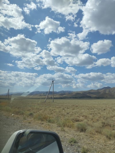The image depicts a vast, dry, grassy plain under a bright blue sky filled with fluffy white clouds. In the foreground, the reflection of a car's side mirror is visible, showing a glimpse of the interior. Two utility poles with wires stretched between them stand in the mid-ground, appearing as solitary structures in the open landscape. In the distance, rolling hills form a hazy backdrop, with a few scattered structures suggesting a small settlement. The overall impression is one of a rural, perhaps arid, environment on a sunny day. The location is identified as the city of Sumbulak, Uzbekistan.