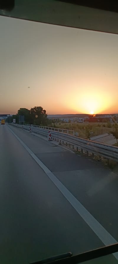 A perspective shot from inside a vehicle shows a highway scene at sunset. The sun is low on the horizon, casting a warm glow across the sky, with hues of orange and yellow dominating the upper portion of the frame. The road ahead is paved and stretches into the distance. A guardrail with red and white reflective markers runs along the right side of the road. Beyond the guardrail, there are trees and a few industrial-looking buildings. The sky is clear except for a few small, dark specks, possibly dust or water droplets on the windshield. The overall impression is of a travel scene during golden hour. The provided hint mentions Sankt Leon, Germany, suggesting this is a highway near that location. There are no people or animals visible in the frame. The scene is static and devoid of any apparent action.