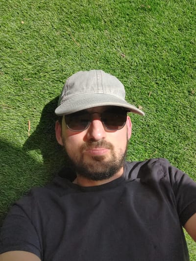 A man lies on a patch of green grass, wearing a dark t-shirt, sunglasses, and a grey cap. The scene is likely outdoors, based on the grass and visible sunlight creating shadows. The location is Havat Shalem, Israel, though this detail is not evident from the image itself.